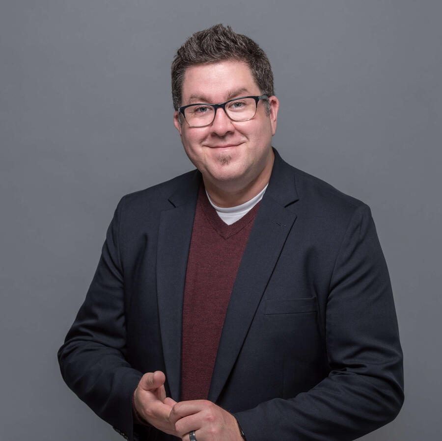 Smiling man in a blazer and maroon sweater stands against a gray backdrop with hands loosely clasped.