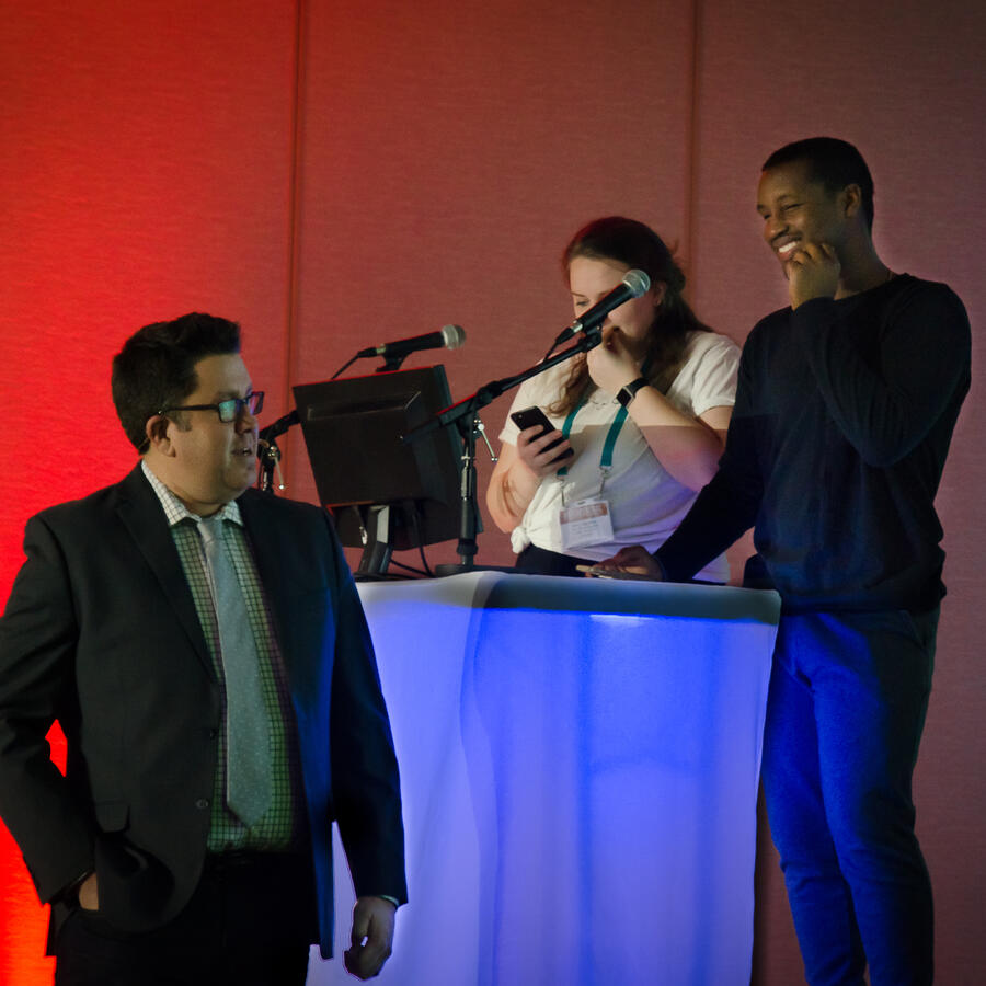 Man in a suit pauses near a lit lectern while two crew members adjust mics and review notes.